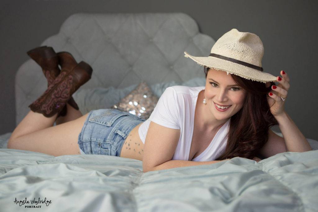 Smiling lady in sexy outfit wearing cowboy hat and cowboy boots in a boudoir session.